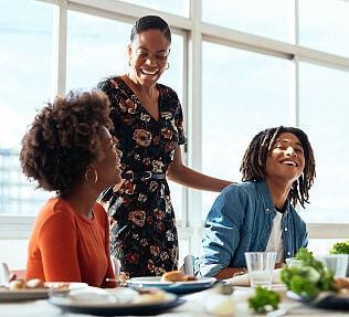 Mother standing behind her adult children as they laugh at the dinner table.