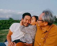 Grandfather watching young granddaughter kiss her father on the temple while sitting outdoors.